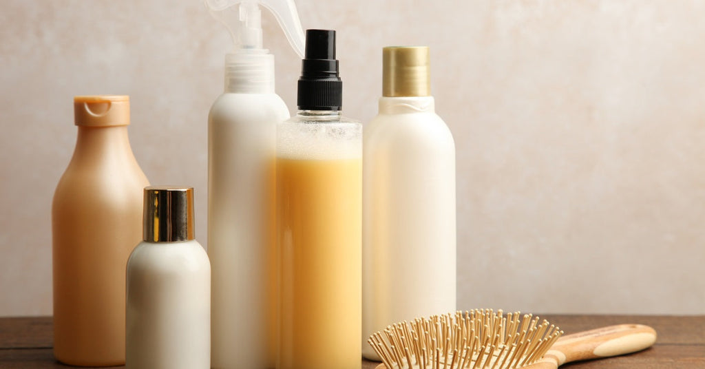 Closeup of five various types of haircare product bottles in different sizes and colors next to a wooden brush.