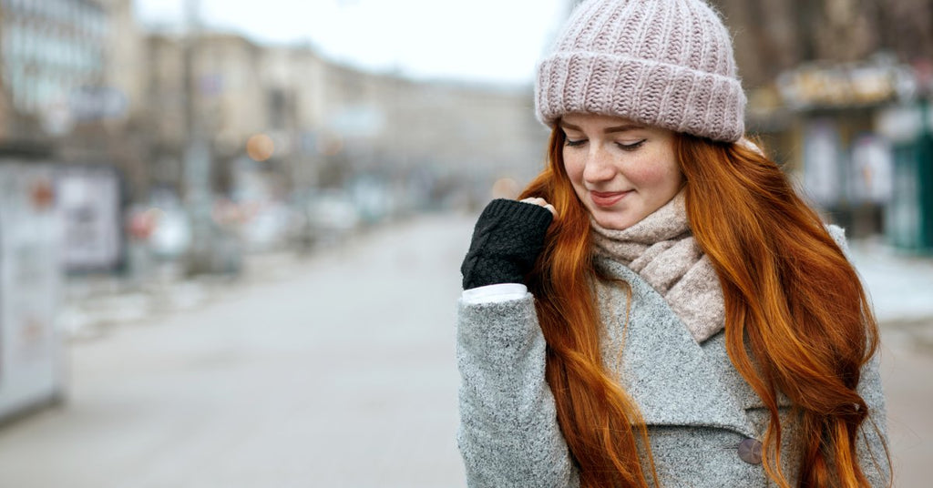 A young woman with long red hair wears a hat, coat, and scarf outside in winter with a blurred background.