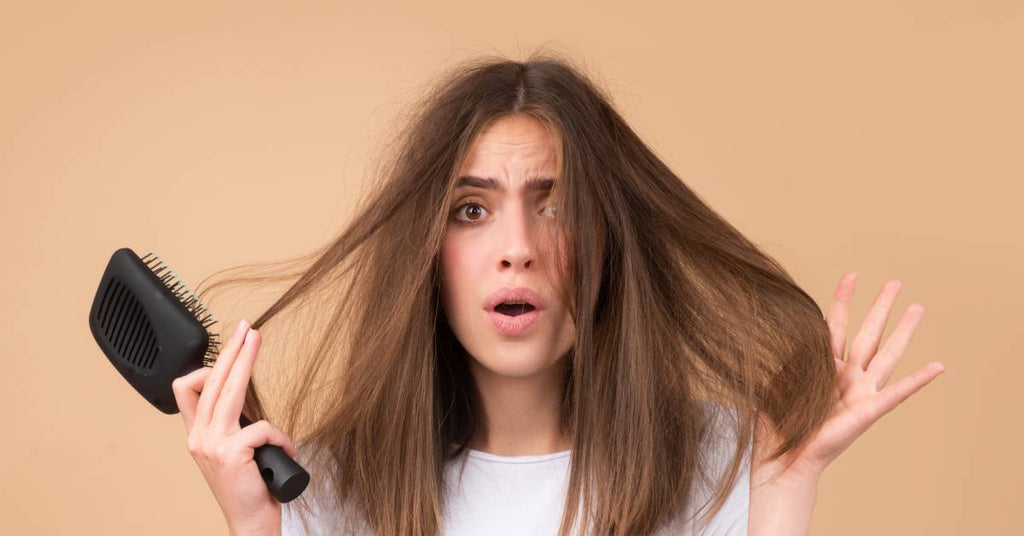 Closeup of a brunette woman looking frustrated while holding a brush in one hand and her frizzy hair in the other.