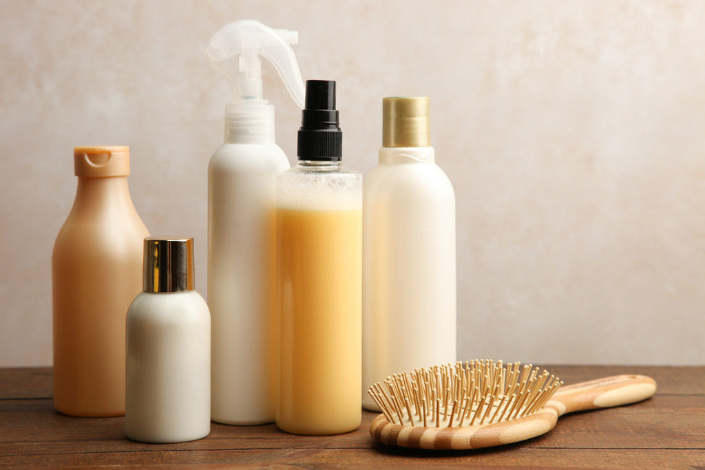 Closeup of five different types of hair care products in various bottles sitting on a table next to a wooden brush.